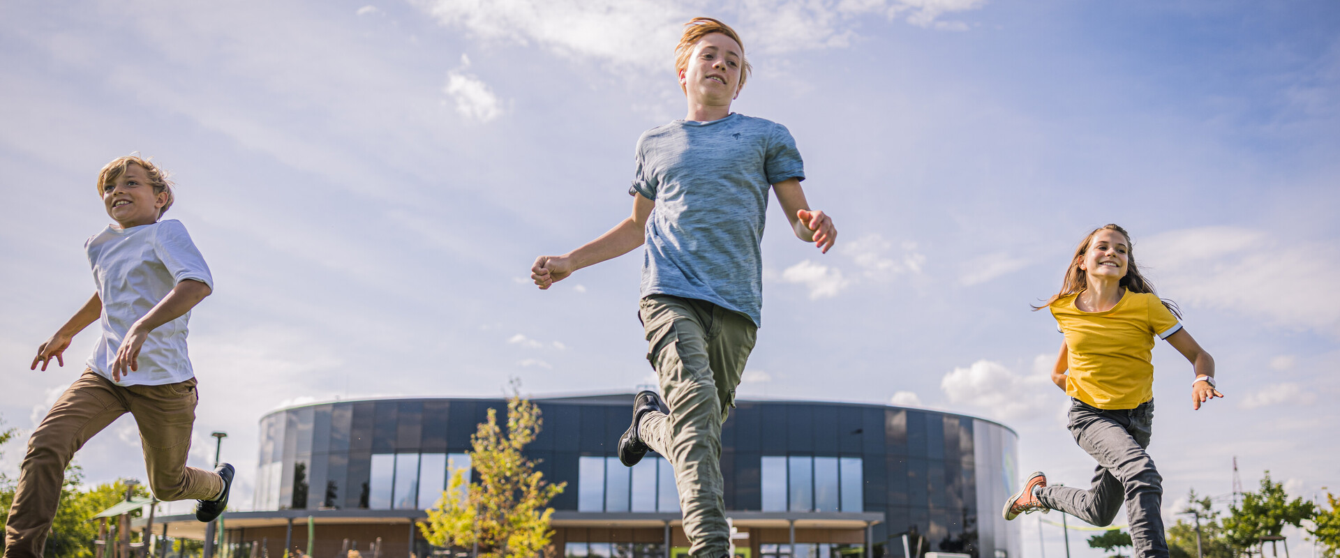 Drei Kinder rennen auf der Wiese im Au&szlig;enbereich, im Hintergrund sieht man das Geb&auml;ude der KLIMA ARENA.